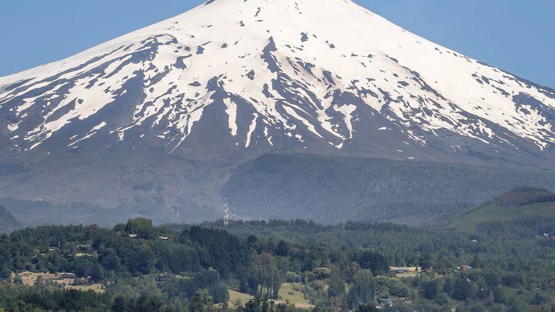 Volcán Villarrica nevado visto desde el lago, con bosque y casas en la ribera